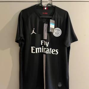 PSG x Jordan svart fotbollströja - Svart PSG fotbollströja i samarbete med Jordan. Klassisk design med vit logga, Fly Emirates-tryck och en smal rand i mitten. Kortärmad och tillverkad i lätt, andningsbart material. Perfekt för dig som vill sticka ut på planen eller på läktaren.