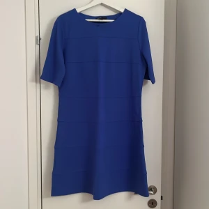 Kornblå kort klänning med kort ärm - En stilren kornblå klänning med kort ärm och rund halsringning. Klänningen har en rak passform och subtila horisontella sömmar som ger en modern look. Perfekt för dig som gillar enkel men snygg design. Den är helt ny oanvänd.