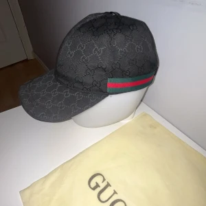 Svart keps från Gucci med GG-mönster - Snygg svart keps från Gucci med klassiskt GG-monogram över hela kepsen. Har grönt och rött band på sidan och justerbar rem baktill i skinn. Perfekt för dig som vill ha en exklusiv och trendig accessoar med streetkänsla.