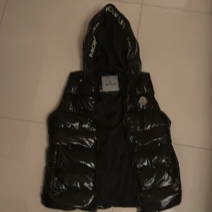 Svart dunväst med huva från Moncler - Snygg svart dunväst från Moncler med glansig finish och stor huva. Västen har dragkedja framtill, två fickor med dragkedja och Moncler-logga på bröstet. Perfekt för lager-på-lager och streetstyle. Västen är inte äkta Moncler & det finns skador på höger sida av västen.