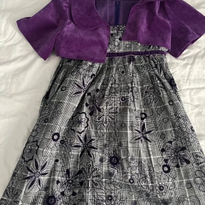 Lila och silver klänning från Papatya - Superfin klänning från Papatya i lila och silver med blommigt och rutigt mönster. Klänningen har en matchande lila bolero, markerad midja och dragkedja bak. Materialet är en mix av bomull och polyester, vilket gör den både mjuk och lätt att bära.