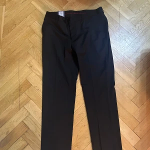 Svarta kostymbyxor Dressmann 31/34 - Klassiska svarta kostymbyxor från Dressmann med raka ben och diskreta bakfickor med knapp. Byxorna har bälteshällor och stängs med knapp och dragkedja. Perfekta för en clean och stilren look.