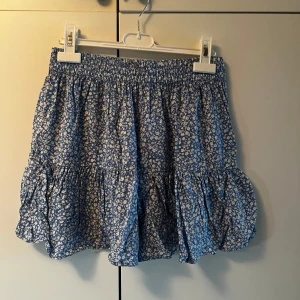 Blommig blå volangkjol från Pigalle - Supersöt blå kortkjol från Pigalle med vitt blommönster och volangdetaljer. Kjolen har resår i midjan för skön passform och är gjord i ett lätt och luftigt bomullstyg. Kjolen har inbyggda shorts! Perfekt för dig som gillar en romantisk och somrig stil! Som man ser på sista bilden saknar den tyvärr snöret man knyter med men det har inte försämrat användningen för mig. Säljer eftersom den är kort på mig. Katt finns i hemmet! 