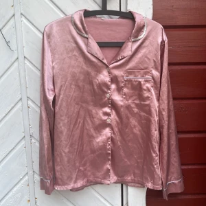 Rosa pyjamas - Säljer en glansig rosa skjorta i satinliknande material med klassisk krage, lång ärm och pärlemorknappar. Skjortan har en bröstficka med vit kant och snygga detaljer vid krage och ärmslut. Perfekt för dig som gillar en mjuk och lyxig känsla.
