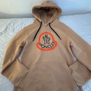 Beige Moncler hoodie  - Snygg beige hoodie från Moncler med stor orange och svart logga framtill och coola färgstänk i blått och rött. Tröjan har huva med svarta snören och en stor magficka. Perfekt för dig som vill sticka ut med en exklusiv streetstyle-look. Storlek:S OBS: PRIS KAN DISKUTERAS!!!