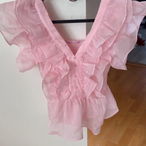 Rosa volangblus med v-ringning - Supersöt rosa blus med v-ringning, volanger längs axlar och framstycke samt knappar framtill. Jag säljer blusen för att det är för liten.Blusen är gjord i ett tunt, transparent material och har korta, luftiga ärmar. Perfekt för dig som gillar romantisk och lekfull stil.