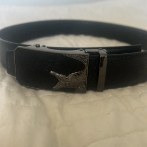 Svart skärp med krokodilspänne - Snyggt svart bälte i skinn med krokodilpräglat mönster och ett coolt metallspänne med krokodilmotiv. Bältet har en stilren och modern look som sticker ut med sin unika detalj på spännet. Perfekt för dig som vill ha något extra till outfiten.