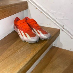 Adidas Crazyfast röda fotbollsskor - Säljer ett par Adidas Crazyfast fotbollsskor i rött med vita detaljer och gula dobbar. Skorna har snörning och är tillverkade i syntetmaterial för lätt känsla och maxad fart på planen. Perfekta för dig som vill sticka ut med stil och speed.