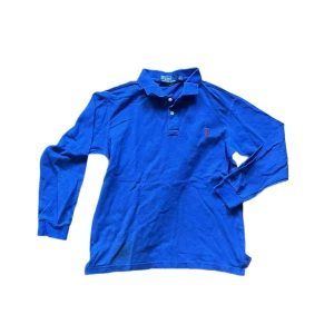 Ralph Lauren piké  - Snygg blå långärmad pikétröja från Polo Ralph Lauren med klassisk krage och knäppning framtill. Tröjan har röd broderad logga på bröstet och är gjord i mjuk bomull. Perfekt för dig som gillar stilren och sportig look. Se missfärgning bredvid måttbandet på bild 4. Jag skickar ej bild när jag bär plaggen och jag vet ej vilken längd och vikt de passar då jag inte prövar kläderna jag säljer 😃