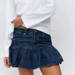 Mörkblå jeanskjol med volang - Supersnygg mörkblå jeanskjol med volangkant och klassiska fickor fram. Kjolen har en kort modell och stängs med knapp och dragkedja. Perfekt för dig som gillar en trendig och avslappnad stil.