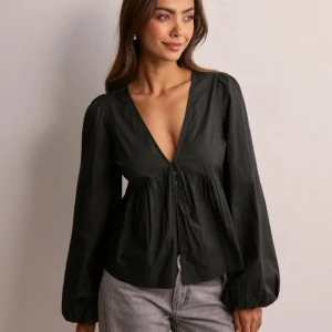 Svart blus med v-ringning och knappar - Svart blus med djup v-ringning och små knappar framtill. Långa puffiga ärmar och lös passform nedtill ger en avslappnad men trendig look. Perfekt att styla med jeans eller kjol för en cool vibe.