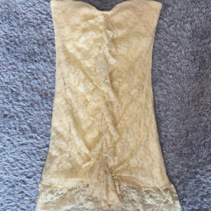Gul spetsklänning med volang - Söt gul axelbandslös kortklänning i spets med volangdetalj framtill. Klänningen har en figurnära passform och är perfekt för dig som vill sticka ut. Spetsen ger en romantisk känsla och volangen framtill gör den extra speciell. Mycket bra skick (lite nopprig men inget som syns när man bär klänningen)💕