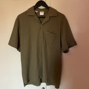 Olivgrön skjorta från Selected Homme - Säljer en olivgrön skjorta från Selected Homme i ribbad bomull. Skjortan har korta ärmar, klassisk krage, bröstficka och knäppning framtill. Perfekt för dig som gillar en avslappnad och stilren look.