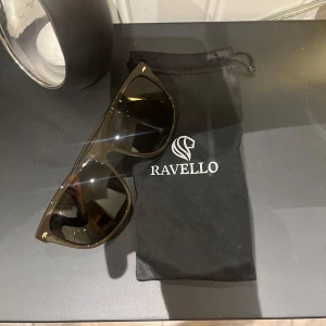 Svarta solglasögon från Ravello - Snygga svarta solglasögon från Ravello med klassisk wayfarer-form. Glasögonen har mörka linser och diskreta guldfärgade detaljer vid bågens hörn. Kommer med en svart tygpåse med Ravello-logga. Perfekta för att lyfta din stil i solen. Ny pris 449kr.