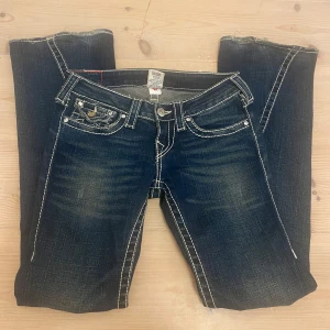 True Religion bootcut jeans mörk blå - Säljer ett par blå jeans från True Religion med tydliga vita kontrastsömmar och ikoniska bakfickor med lock och knapp. Modellen har bootcut-ben och låg midja. Klassisk denim med snygga detaljer och patch bak i midjan. Den saknar en pärla på fram och bak knappen men man tänker inte på det ( man ser på bilderna) hör av er vid frågor❤️❤️