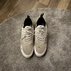 Nike Air Max 270 vita sneakers - Nike Air Max 270 sneakers i vitt med svarta detaljer och synlig Air Max-dämpning i hälen. Ovandelen är i ventilerande syntetmaterial med mesh och snörning framtill. Snygg sportig look med rund tå och platt sula. Perfekta för dig som gillar streetstyle.