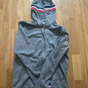 Grå Moncler hoodie - Snygg grå hoodie från Moncler med hel dragkedja och huva. Tröjan har Moncler-logga på ärmen och coola ränder i rött, vitt och blått på huvan. Perfekt för dig som gillar stilrena och sportiga vibes. Skriv om ni har några frågor eller funderingar!