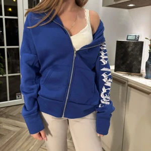 Blå hoodie med vit text på ärmen - Snygg blå hoodie med dragkedja och huva. Ärmen har en cool vit text i grafisk stil som ger streetkänsla. Hoodien är fodrad med mjuk fleece på insidan och har en relaxed passform. Perfekt för chill dagar eller när du vill ha en avslappnad look.