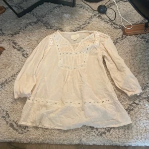 Beige blus från H&M Conscious Collection - Romantisk vit blus från H&M Conscious Collection med v-ringning, broderade spetsdetaljer och diskret prickstruktur i tyget. Långärmad modell med lös passform och fina veck framtill. Perfekt för dig som gillar boho-vibes och detaljer.