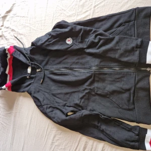 Svart hoodiejacka med dragkedja - Svart hoodiejacka från Moncler med hel dragkedja, huva med snörning och vita samt röda detaljer på huvan och ärmsluten. Jackan har två fickor framtill och en broderad Moncler-logga på bröstet. Knappt använd, som ny.