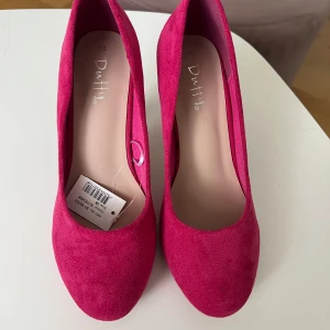 Ceriserosa pumps från Duffy storlek 38 - Snygga ceriserosa pumps från Duffy i mockaimitation. Skorna har rund tå, hög klack och en klassisk siluett som verkligen sticker ut. Perfekta för dig som vill addera färg till din outfit. Storlek 38.