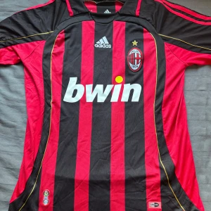 AC Milan randig matchtröja Adidas - Säljer en klassisk AC Milan fotbollströja från Adidas med röda och svarta vertikala ränder, klubbmärke och Bwin-tryck på bröstet. Tröjan har korta ärmar, gulddetaljer och är tillverkad i lätt och ventilerande ClimaCool-material.