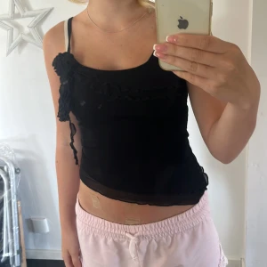 Svart linnetopp med volangdetaljer - Säljer en svart, tight linnetopp med volangdetaljer framtill. Toppen har en dekorativ rosett på ena axeln och är lätt transparent med en croppad passform. Perfekt för dig som gillar trendiga och feminina detaljer. 