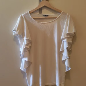 Vit volangblus från Ellos - Supersöt vit blus från Ellos med volangärmar och lätt ribbad struktur. Blusen har rund halsringning och flowiga korta ärmar med flera lager volanger som ger en extra touch. Perfekt för dig som gillar en feminin och luftig stil.