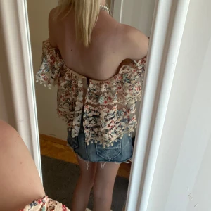 Blommig offshoulder blus med spets - Superfin blommig offshoulder blus i beige spets med små färgglada blommor. Toppen är croppad, har volangiga ärmar och söta bollar längs kanterna. Dragkedja i ryggen som ger en extra snygg touch. Perfekt för sommarens alla häng!