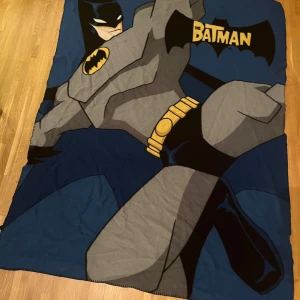 The Batman - Den här filten visar en stor, färgstark illustration av Batman i klassisk stil. Perfekt för fans av superhjältar och serietidningar som vill ha något unikt och coolt i sitt rum. Motivet är tydligt och passar både unga och unga vuxna som gillar DC Comics.
