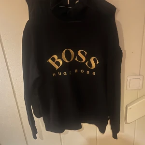 Svart sweatshirt från Hugo Boss - Svart sweatshirt från Hugo Boss med stort guldigt BOSS-tryck på bröstet. Klassisk rund halsringning och långa ärmar. Perfekt för dig som gillar stilrena och trendiga plagg med tydlig logga.