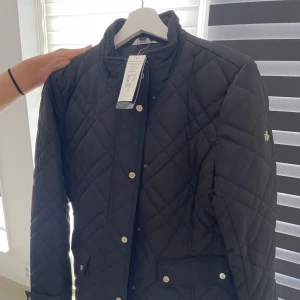 Svart quiltad jacka från Ralph Lauren - HELT NY - Snygg svart quiltad jacka från Ralph Lauren med RL-logga på ärmen. Jackan har guldiga knappar, ståkrage och två fickor framtill. Perfekt för dig som vill ha en stilren och klassisk look under höst och vår. Etiketten är fortfarande kvar, dvs den är helt oanvänd! 🙌