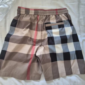Burberry shorts - Snygga shorts från Burberry med klassiskt rutigt mönster i beige, svart, blått och rött. De har elastisk midja med svart snörning och praktiska fickor. Perfekta för sommaren och ger en trendig vibe till din outfit.