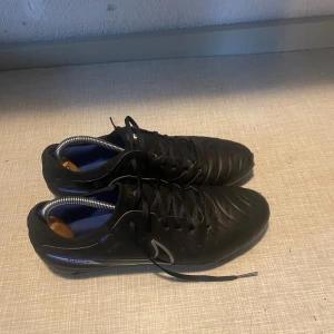 Nike Tiempo svarta fotbollsskor - Svarta Nike Tiempo fotbollsskor med blå detaljer och silverfärgad sula. Skorna har snörning, rund tå och är tillverkade i skinn. Perfekta för dig som vill ha stilrena och klassiska fotbollsskor med bra grepp på planen.