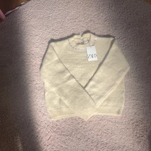 Ljusgul/beige stickad tröja med pärlor Zara - Superfin ljusgul/beige stickad tröja från Zara med pärldetaljer runt halsen. Tröjan har lång ärm och ribbade muddar vid ärmslut och nederkant. Perfekt för dig som vill ha en söt och trendig look med lite extra bling.