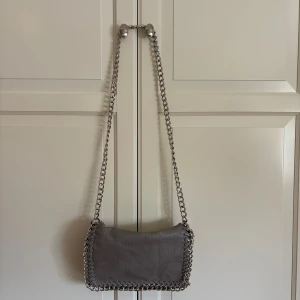 Grå axelväska med kedja från Tiamo - Snygg grå axelväska från Tiamo med grov silverfärgad kedja som axelrem och dekorativ kedjedetalj runt kanterna. Väskan är tillverkad i skinnimitation och har dragkedja baktill för extra förvaring. Perfekt för dig som gillar stilrena accessoarer med edge.