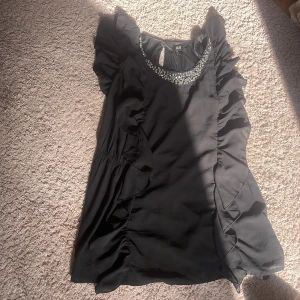 Svart klänning med volanger från H&M - Supersnygg svart klänning från H&M med glittrande paljetter och stenar runt halsringningen. Klänningen har korta volangärmar och volangdetaljer längs sidorna. Perfekt för dig som gillar statement-plagg med extra bling och coola detaljer.