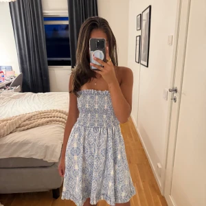 Blåvit kort klänning från Salant - Supersöt blåvit kort klänning från Salant med smockad överdel och axelbandslös design. Klänningen har ett fint mönster i blått och vitt, volangkant nertill och är perfekt för varma dagar. Lätt och luftig känsla, passar dig som gillar somriga vibes.💞