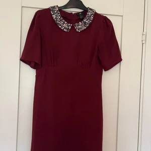 Vinröd klänning med pärlkrage H&M - Supersnygg vinröd kortklänning från H&M med korta ärmar och en statementkrage täckt av glittrande pärlor och stenar. Klänningen har en klassisk rak passform och dragkedja bak. Perfekt för dig som vill sticka ut med en cool detalj.