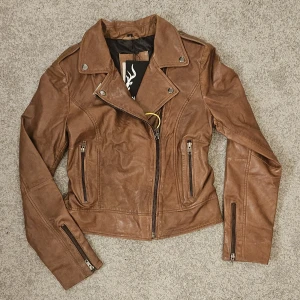 Helt ny bikerjacka i äkta läder – NOTYZ, stl 40 - Säljer en exklusiv bikerjacka i 100% äkta läder från NOTYZ – helt ny, med prislapp kvar! Färgen är en varm cognac som lyfter varje outfit – tidlös, snygg och trendig på samma gång.  * Detaljer som sticker ut: * - Asymmetrisk dragkedja framtill  - Flera praktiska fickor med dragkedjor  - Tryckknappar på kragen - Dragkedjor vid ärmslut för extra edge  * Storlek: 40 (normal passform)  * Skick: Helt ny – aldrig använd  * Nypris: 2 549 kr  En statement-jacka som tar din stil till nästa nivå – perfekt
