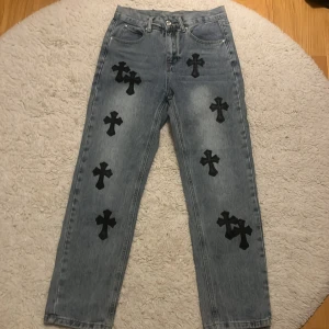 Chrome Hearts jeans med svarta kors - Unika ljusblå jeans från Chrome Hearts med flera svarta korsapplikationer på både fram- och baksida. Klassisk femficksmodell med silverdetaljer och svart läderpatch bak. Raka ben och cool streetwear vibe. Perfekta för dig som vill sticka ut.