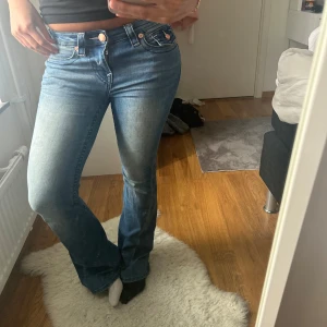 True Religion blå bootcut jeans - Säljer ett par blå jeans från True Religion med bootcut passform. Jeansen har klassiska kontrastsömmar, snygga fickdetaljer med lock och knappar bak samt ikonisk patch i midjan. Perfekta för dig som gillar en avslappnad men trendig look.