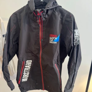 Svart vindjacka från Pelle P x Stena Match Cup - Svart vindjacka med röda dragkedjor och vita tryckdetaljer från Pelle P x Stena Match Cup Sweden. Jackan har huva, meshfoder och coola tryck på både bröst, ärm och rygg. Perfekt för dig som gillar segling eller vill ha en sportig look.