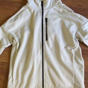 Vit Flood Zip Hood från Sail Racing - Snygg vit Flood Zip Hood från Sail Racing med hel dragkedja och huva. Jackan har en bröstficka med dragkedja och diskret logga. Tillverkad i en skön mix av bomull och polyester, perfekt för chill vår- och höstdagar. Endast använd 1 gång