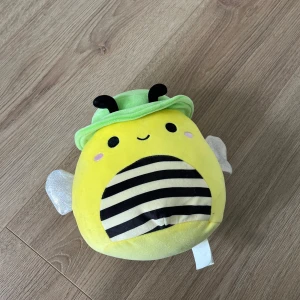 Squishmallow  - Bilderna visar en söt gosedjursbi från Squishmallows med grönt hatt, glittriga vingar och ett glatt ansikte. Den är mjuk, färgglad och perfekt för att mysa med eller dekorera rummet. Passar dig som gillar gulliga och trendiga mjukdjur.