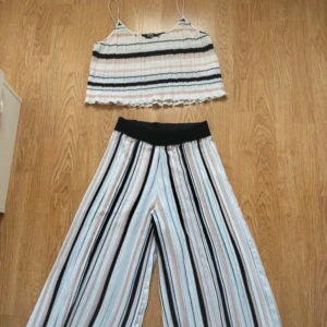 Randigt set - Säljer ett par luftiga wide byxor från Bik Bok med svart resår i midjan och tillhörande linne. Setet har randigt mönster i vitt, svart och rosa samt är tillverkade i ett lätt, plisserat tyg. Perfekta för en chill och trendig look