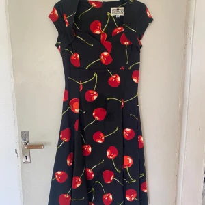 Svart klänning med körsbär från Collectif - Supersöt svart klänning från Collectif med röda körsbär och gröna stjälkar över hela tyget. Klänningen har korta ärmar, v-ringning och en utställd kjol som ger en retrovibe. Perfekt för dig som gillar färgglada och lekfulla prints!