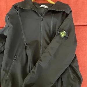Svart vindjacka från Stone Island - Svart vindjacka från Stone Island med ikonisk patch på ärmen. Jackan har huva, flera dragkedjor framtill och snedställda fickor. Tillverkad i lätt och slitstark polyester, perfekt för dig som gillar streetwear och snygg design.