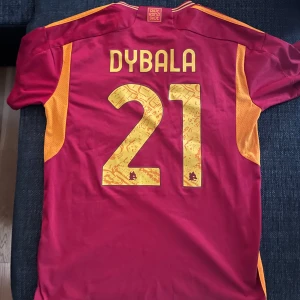 AS Roma Dybala 21 matchtröja Adidas M - AS Roma fotbollströja med Dybala 21 på ryggen. Röd med gula detaljer, klassiska Adidas-ränder på axlarna och klubbens logga på bröstet. Tillverkad i lätt och ventilerande Aeroready-material, perfekt för match eller träning.