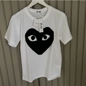 Vit t-shirt Comme des Garçons Play - Vit t-shirt från Comme des Garçons Play med stort svart hjärta och ögon-tryck på bröstet. Klassisk rund halsringning och korta ärmar. Tillverkad i mjuk bomull, perfekt för en clean och cool stil.
